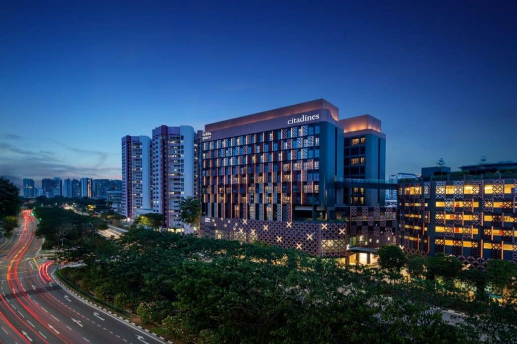Fachada do hotel Citadines Rochor, uma das recomendações de onde ficar em Singapura. A foto foi tirada à noite e há luzes acesas nas janelas do quarto do hotel. Há outros prédios ao redor. Na frente há várias árvores que separam os prédios de uma avenida da cidade.