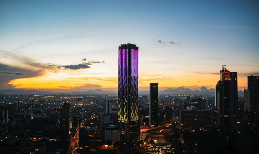 Arranha-céu iluminado por luzes roxas e amarelas ao pôr do sol para ilustrar o post sobre chip de celular para Bogotá. Ao redor há outros prédios e casas iluminadas. - Foto: Santiago Boada via Pexels