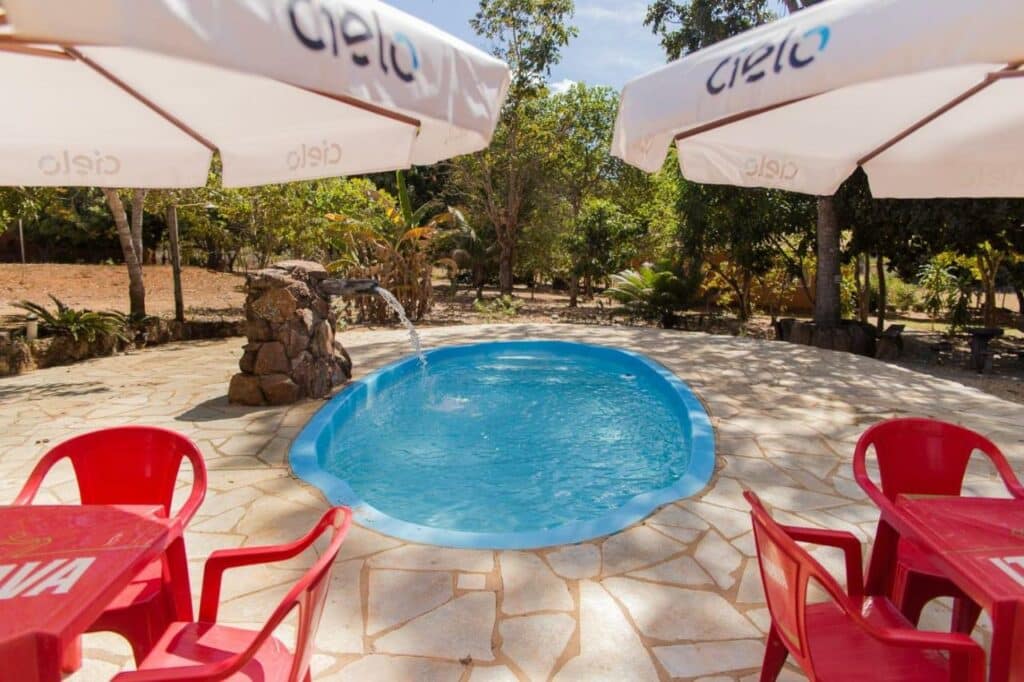 Área externa da piscina no Condomínio Shambala Piri, localizado em Pirinópolis. Piscina com cascata ao centro, ao redor cadeiras e mesas e guarda sol, ao fundo vasta vegetação.