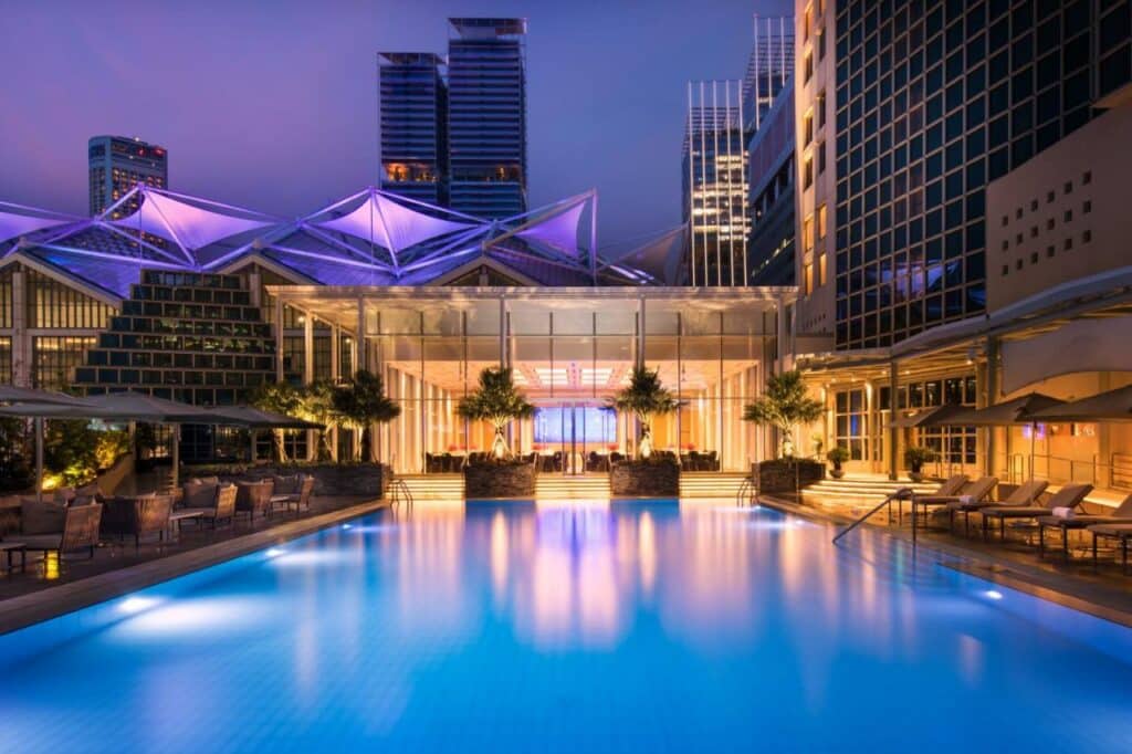 Piscina do hotel Conrad Centennial Singapore. Há diversas cadeiras e espreguiçadeiras bege nas laterais da piscina, e algumas árvores pequenas de frente para a entrada do hotel. Ao fundi podemos ver alguns prédios.