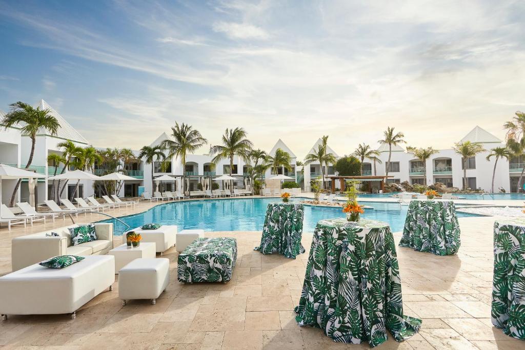 área da piscina do Courtyard by Marriott Aruba Resort com sofás brancos e mesas cobertas com toalhas de mesa estampadas de folhas de palmeiras