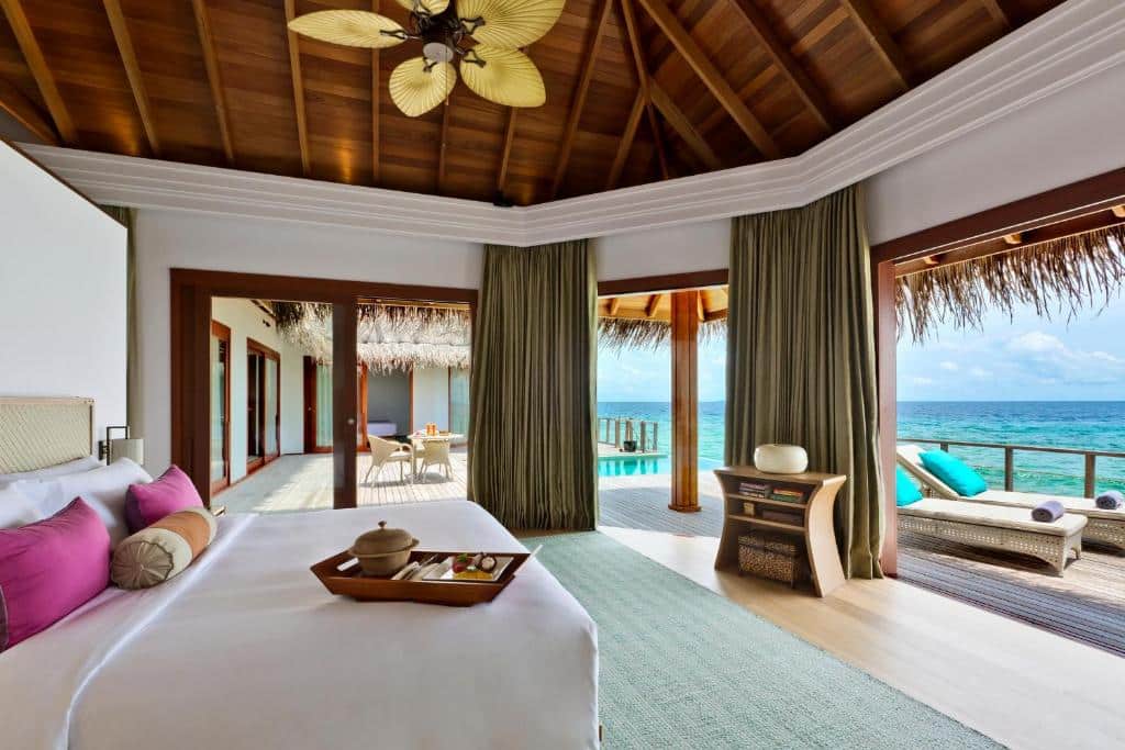 Quarto do Dusit Thani Maldives, com cama de casal, uma bandeja com alimentos em cima e vista panorâmica do mar cristalino de Maldivas. Há um deck de madeira com espreguiçadeiras e uma piscina de borda infinita