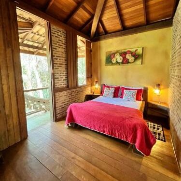 Bangalô da Eco Pousada VerDivino, com chão de maeira, paredes de tijolinhos à vista e uma cama de casal. O ambiente é charmoso e rústico, há uma rede na varanda com vista para a natureza para ilustrar as pousadas em Macacos