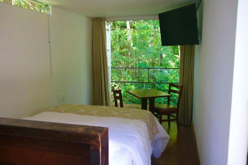 Interior de um dos Econtainer Suítes Macacos, de 17 m², com uma cama de casal, uma mesa com duas cadeiras, uma TV e uma porta de vidro com varanda e vista para a natureza verde