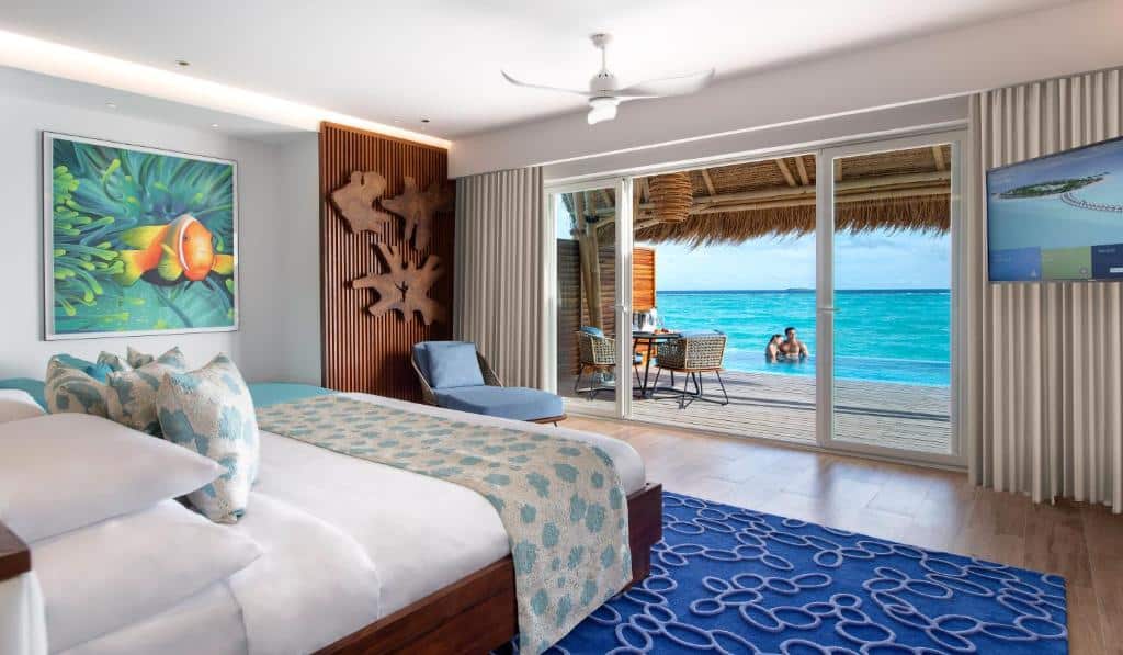 Vila sobre a água com piscina, de 187 m², do Emerald Maldives Resort, com cama de casal, uma poltrona azul e varanda com piscina de borda infinita (tem um casal abraçados dentro) e o mar ao lado azulado. Imagem para ilustrar o post de hotéis em Maldivas