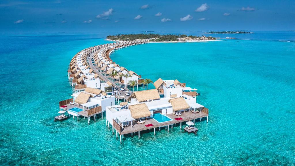 Avenida em cima do mar com bangalôs submersos em um mar azul cristalino. Ao fundo, há uma ilha que faz parte do Emerald Maldives Resort, com bastante natureza e praia privativa.