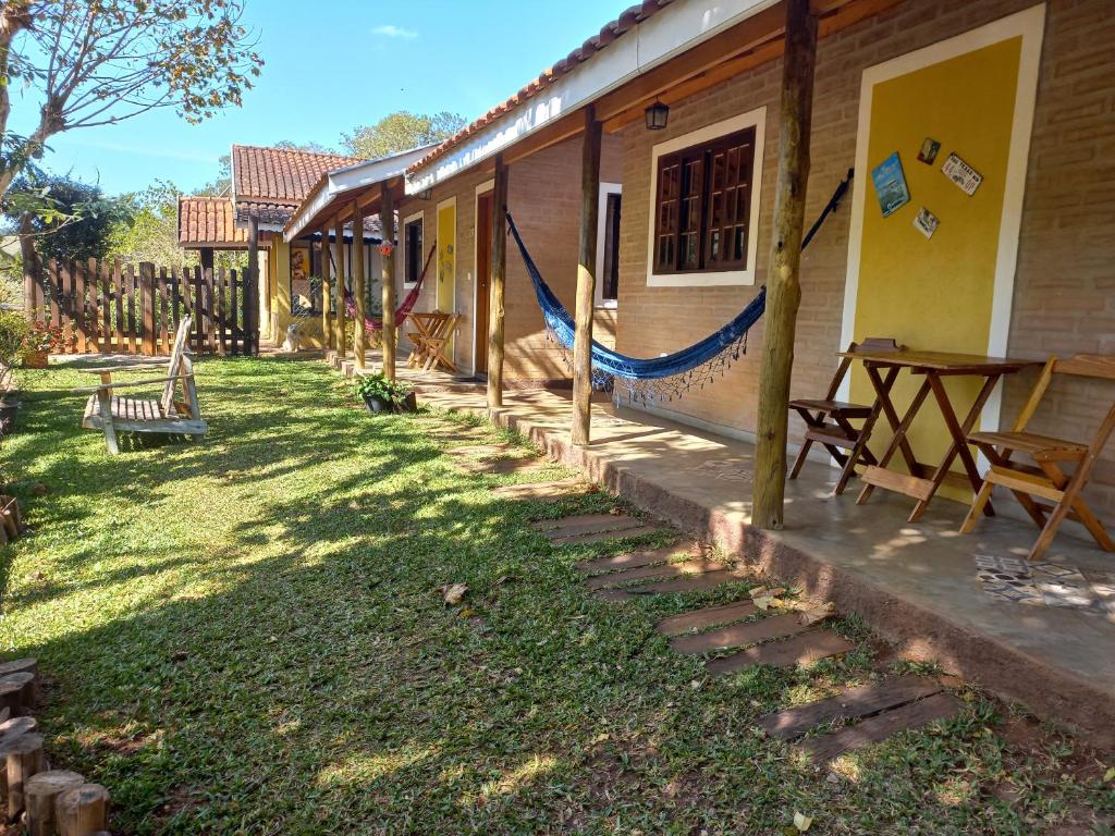 Frente do Chalés do Alcantilado durante o dia, com estrutura de tijolinho, portas amarelas, mesa de madeira com duas cadeiras, rede, com um gramado na frente. Representa chalés em Santo Antônio do Pinhal
