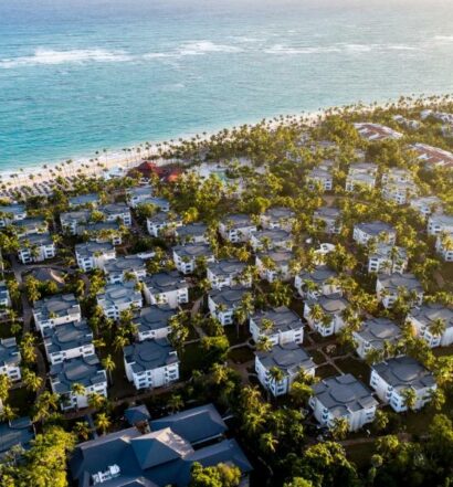 Vários prédios espalhados com árvores espalhadas em volta e no meio, em uma área beira-mar, com areia branca e mar azulado. Imagem para ilustrar o post como escolher um resort em Punta Cana