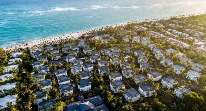 Vários prédios espalhados com árvores espalhadas em volta e no meio, em uma área beira-mar, com areia branca e mar azulado. Imagem para ilustrar o post como escolher um resort em Punta Cana