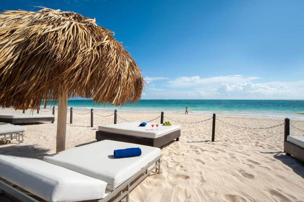 Espreguiçadeiras e camas espalhadas em uma área cercada à beira-mar, com areia branquinha e mar azulado cristalino