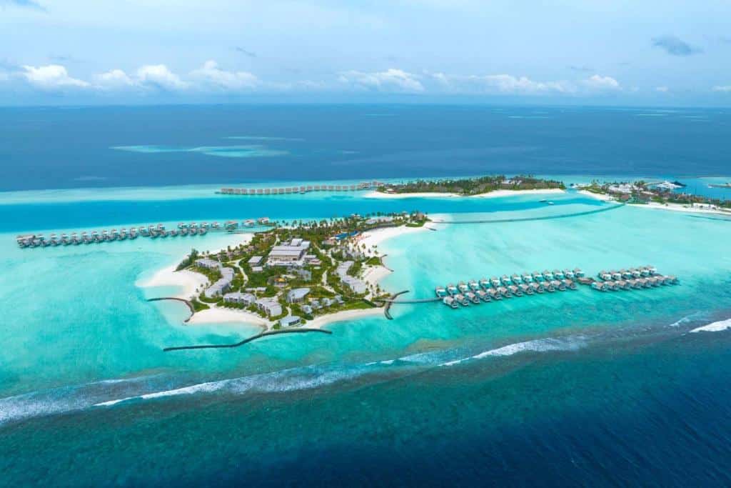 Vista aérea do Hard Rock Hotel, em Maldivas, com três ilhas privativas com os prédios e estrutura do hotel e alguns bangalôs flutuantes no mar num tom azulado cristalino