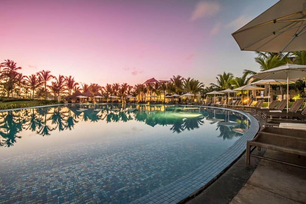 Piscina do Hard Rock Hotel & Casino, com o céu num tom degradê lilás e azul refletindo na piscina, com espreguiçadeiras em volta, árvores e luzes acesas de um prédio do hotel. Imagem para ilustrar os resorts em Punta Cana