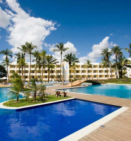 Amplas piscinas em tons de azul escuro e claro com pequenas ilhotas com palmeiras e, mais ao fundo, há o hotel, para representar hotéis em São Luís do Maranhão