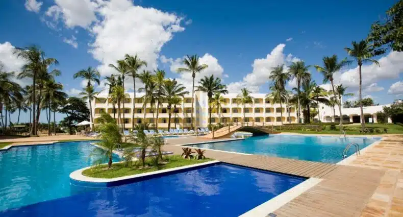 Amplas piscinas em tons de azul escuro e claro com pequenas ilhotas com palmeiras e, mais ao fundo, há o Blue Tree Towers São Luís, para representar hotéis em São Luís do Maranhão