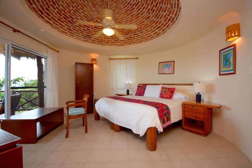 Quarto no Hotel La Joya Isla Mujeres com uma cama de casal, ventilador de teto, uma sacada com cortina, duas mesinhas de cabeceira e alguns quadros nas paredes, para representar hotéis em Isla Mujeres