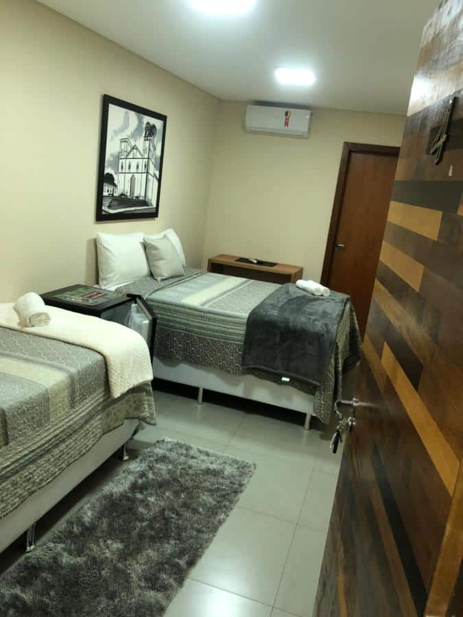 Quarto com uma cama de casal e outra cama de solteiro em Hotel Santa Maria em Pirenópolis. Quarto possui ar condicionado, mesa de cabeceira, quaro pendurado na parede e frigobar.