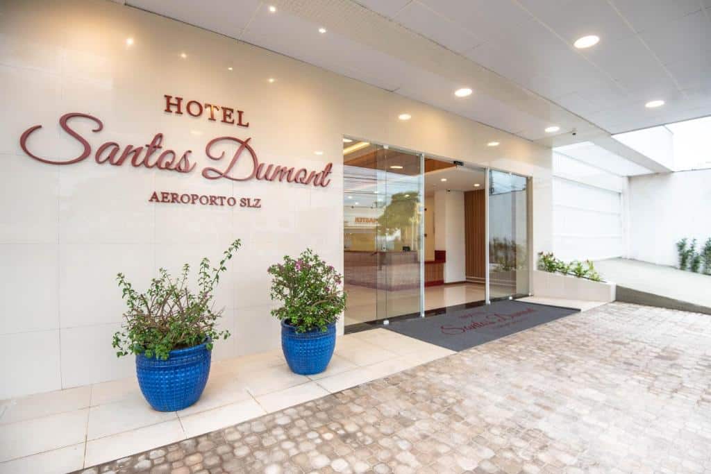 Entrada do Hotel Santos Dumont Aeroporto SLZ com portas de vidro, iluminação indireta e dois vasos de plantas, para representar hotéis em São Luís do Maranhão