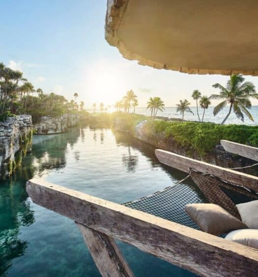 Uma rede suspensa cm almofadas de frente para uma enorme piscina no Hotel Xcaret Arte - All Parks All Fun Inclusive - Adults Only com alguns coqueiros ao redor, para representar hotéis em Playa del Carmen