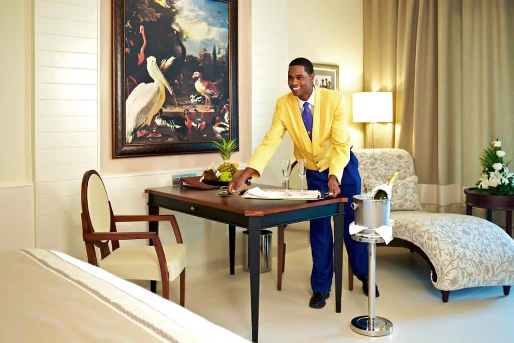 Homem negro vestido de um terno azul e amarelo arrumando a mesa com duas taças em cima e uma bandeja de frutas ao lado. Ao lado da mesa há um balde com um espumante dentro e uma poltrona ao lado para ilustrar como escolher um resort em Punta Cana