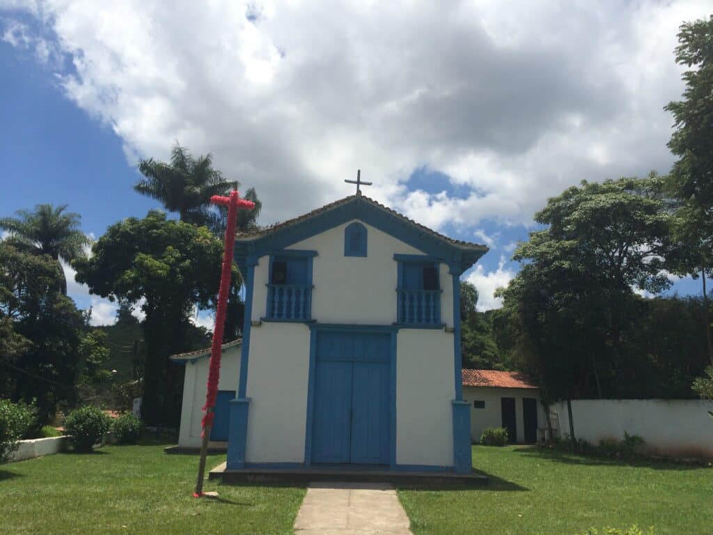 Igreja pequena com parede azul, sendo as bordas do edifício, janelas e porta azuis, com uma cruz no meio e em cima. Há um gramado verde em volta e muita natureza nas redondezas