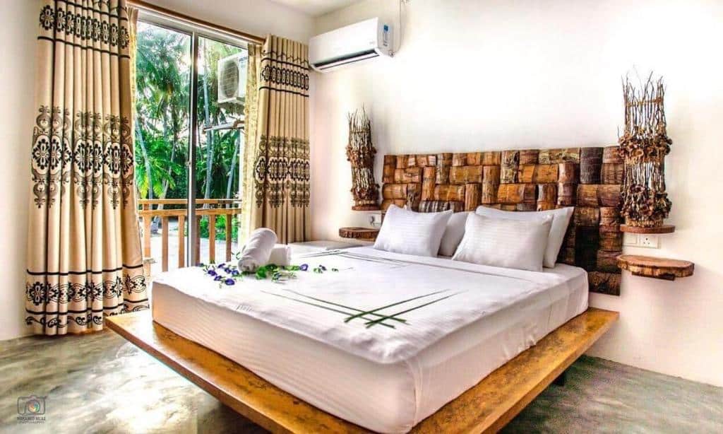 Quarto do Island Break, um dos hotéis em Maldivas, com decoração rústica, sendo a cama de casal com base de madeira e a cabeceira artesanal composta por vários pedaços de troncos de árvore. O quarto tem ar-condicionado e porta de vidro mostrando a natureza ao redor
