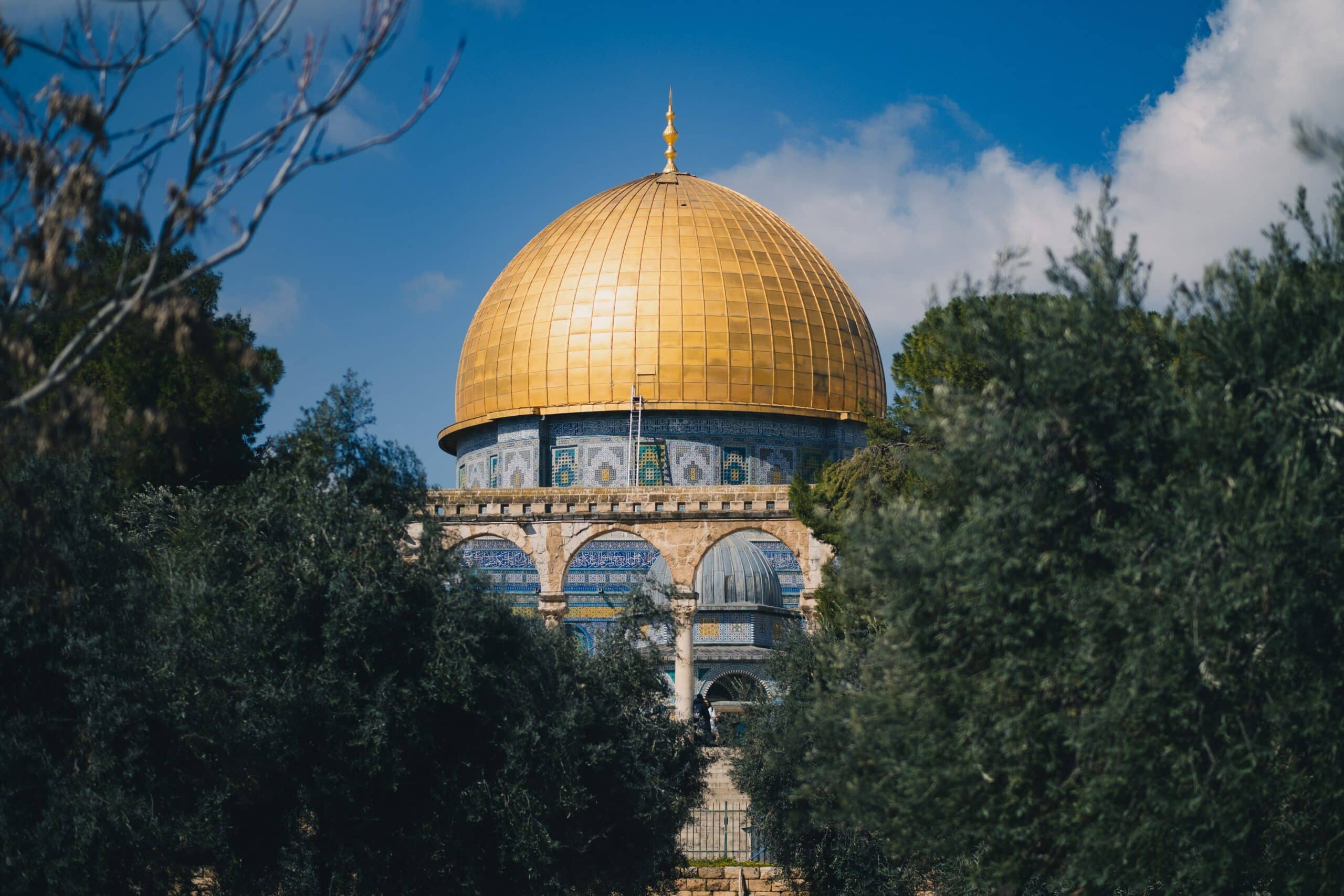 Vista da Cúpula da Rocha, Jerusalém durante o dia com cúpula dourada, paredes azuis e árvores na frente. Representa chip celular Jerusalém.