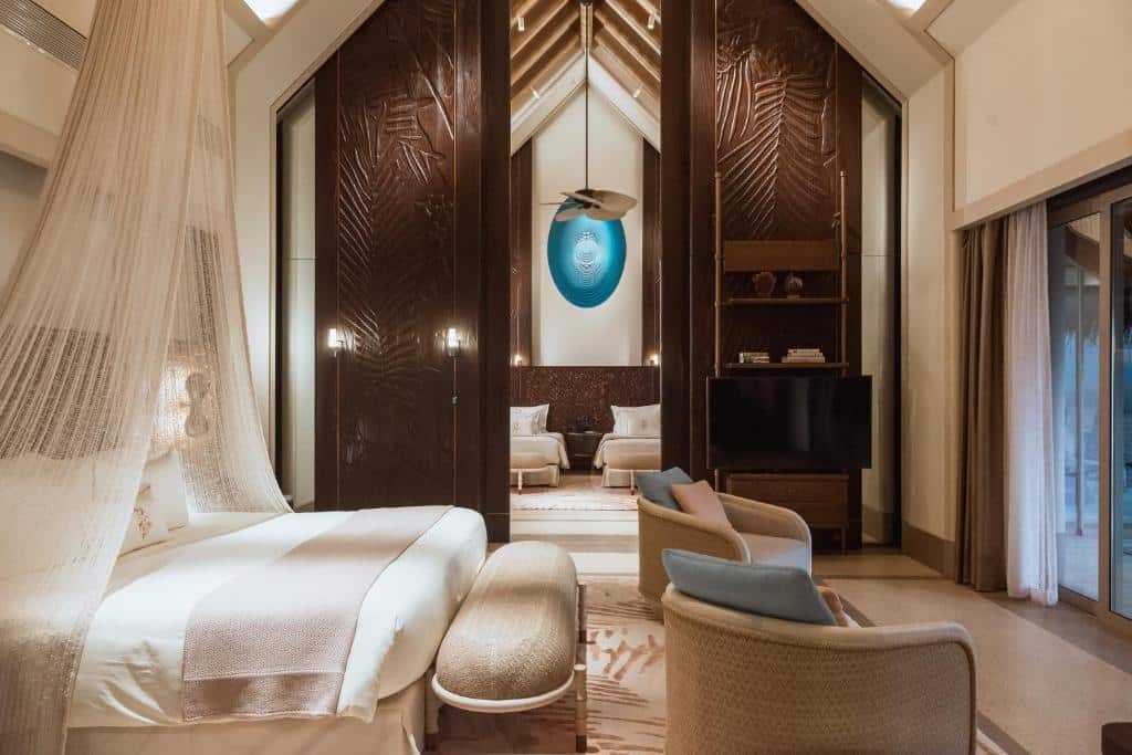 Vila do Joali, um dos hotéis em Maldivas, de 280 m², com cama de casal, duas poltronas na frente e paredes altas de madeira com a porta aberta ao meio mostrando duas camas de solteiro em outro quarto. Há uma TV e portas de vidro dando acesso à varanda