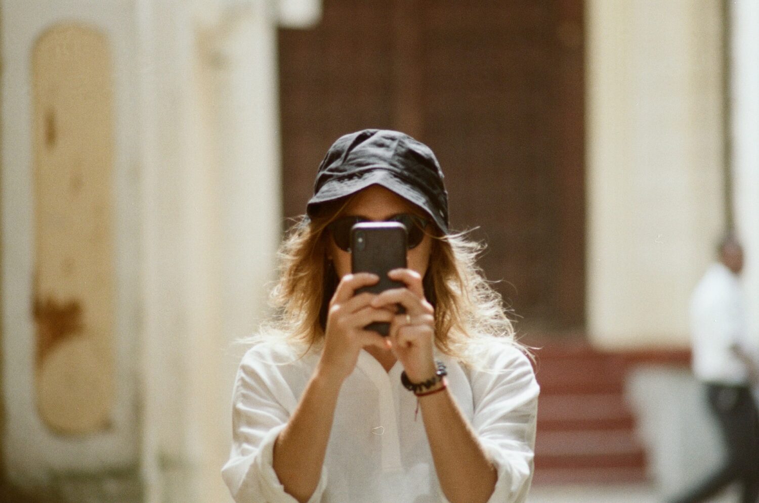 Uma mulher branca de cabelo longos em tons de castanho claro, com um chapéu preto, uma camisa de botões branco, segurando um celular na vertical, para representar chip celular Zanzibar
