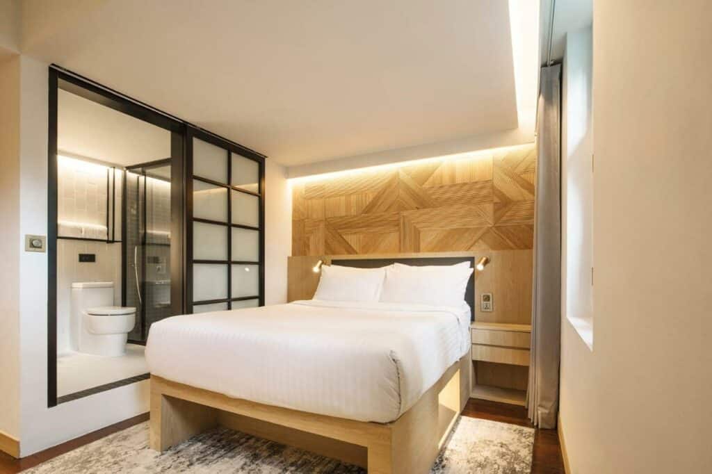 Quarto do hotel KēSa House, The Unlimited Collection by Oakwood, uma das recomendações de onde ficar em Singapura. A cama de casal é de madeira com jogo de cama branco. Há uma mesinha de cabeceira de cada lado da cama. Há uma janela na parede ao lado da cama com uma cortina bege, e um tapete embaixo da cama.
Do lado direito podemos ver o banheiro, que é separado do quarto por uma porta de correr. A privada é branca e ao seu lado está um box transparente com chuveiro.