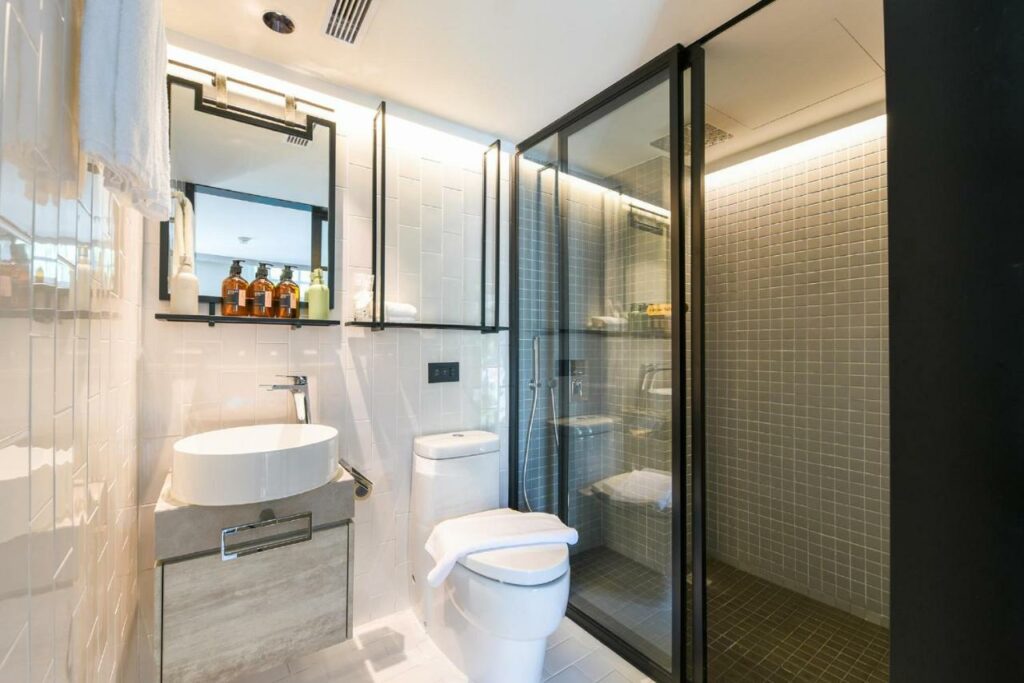 Banheiro do hotel KēSa House, The Unlimited Collection by Oakwood. A privada branca está entre uma pia  bege e o box transparente com chuveiro. Na parede acima da pia há um suporte com espelho e produtos de banho.