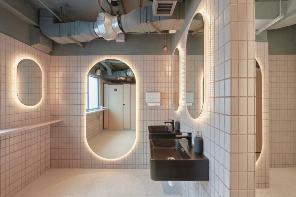 Banheiro do KINN Capsule Hotel. Há diversos espelhos iluminados nas paredes de ladrilhos bege. O espelho da frente reflete a imagem de uma porta de banheiro fechada. Na parede da direita há pias pretas com frascos de sabonete. Um porta-papel está no fim do cômodo.