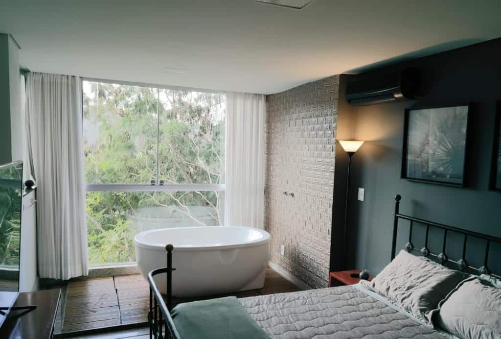 Loft do Loft Espaço Vila da Serra, de 65 m², com uma cama de casal e uma banheira ao lado de uma parede de vidro com vista da natureza verde. Há também uma escrivaninha com uma TV em cima para ilustrar as pousadas em Macacos