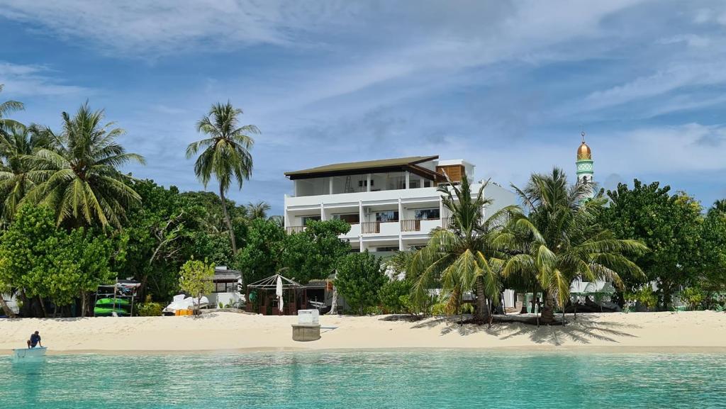 Praia onde fica o Luau Beach Inn, com um barco pequeno no mar com um moço em cima, areia branca, várias árvores na areia, com o prédio branco do hotel atrás. Imagem para ilustrar o post de hotéis nas Maldivas