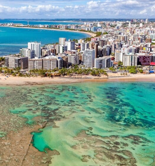 Costa do mar de Maceió, com um mar transparente mostrando barreiras de corais, areia branca e prédios ao meio.