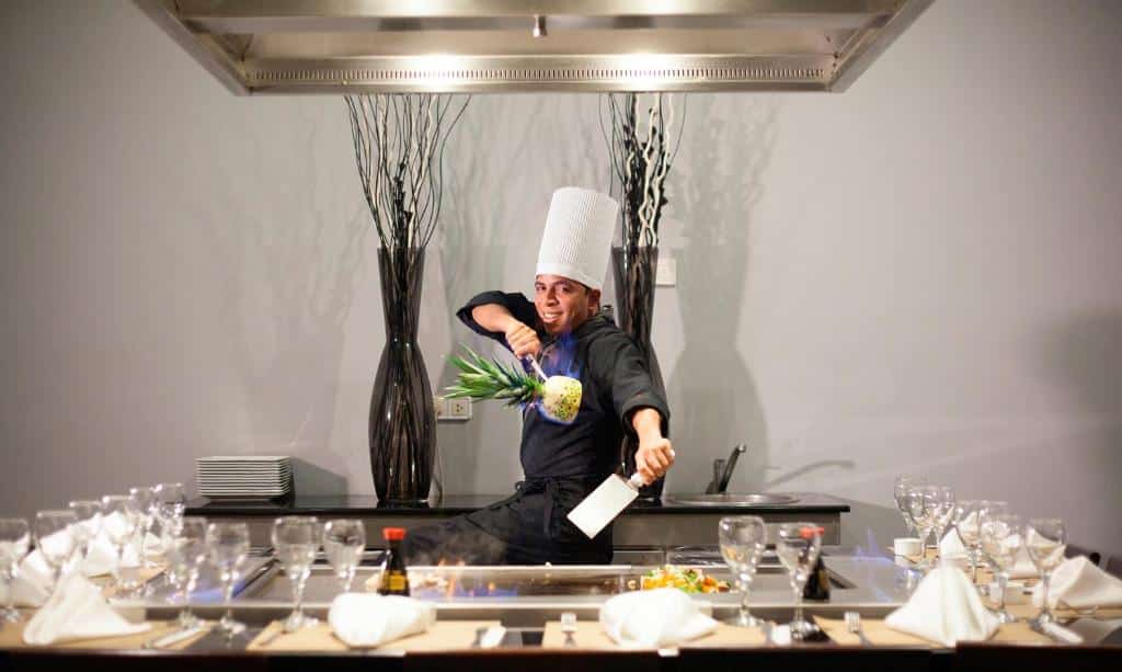 Chefe de cozinha do Majestic Elegance Punta Cana usando roupa preta, chapéu branco, e segurando com uma mão uma espátula e na outra um abacaxi. Em volta dele há uma mesa desfocada com taças e guardanapos espalhados em cima