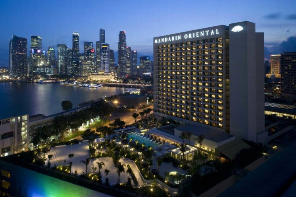 Visão da fachada do hotel Mandarim Oriental Singapura. A foto foi tirada ao anoitecer, e o hotel está iluminado. Na frente do hotel, há uma piscina luxuosa cercada por palmeiras. Ao fundo há diversos prédiostambém iluminados.
