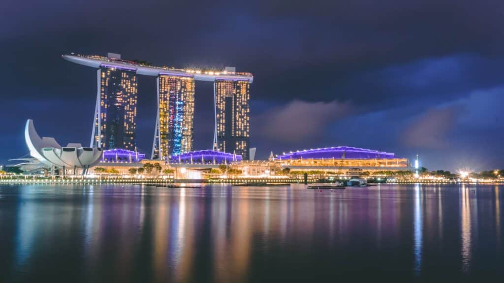 Foto do Marina Bay Sands à noite. O hotel está todo iluminado e suas luzes refletem no mar em frente. Há outras construções iluminadas ao redor também. - Foto: Harish Shivaraman via Unsplash