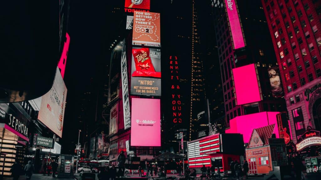 vista da Times Square em Nova York a noite, com diversões telões coloridos em diferentes prédios. A cor rosa, no entanto, destoa das demais, uma vez que aparece em vários telões fazendo menção a marca T-Mobile