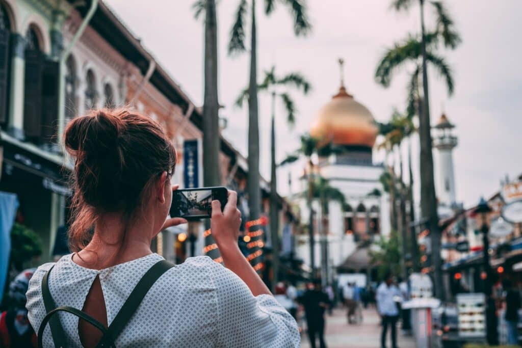 pessoa de costas segurando um celular tirando uma foto Mesquita do Sultão, em Rochor, Singapura, um dos países que pode ser visitada com o chip Vivo no exterior, o fundo está meio desfocado e há palmeiras e pessoas andando pela rua em frente à mesquita