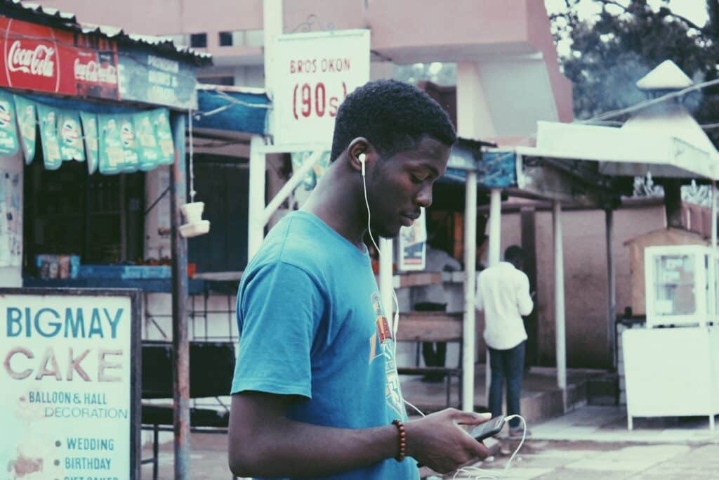 Um homem negro de lado, usando uma camiseta azul enquanto segura um celular com o fone conectado no aparelho e em seu ouvido, ao redor há algum comércio local na cidade de Kaduna na Nigéria