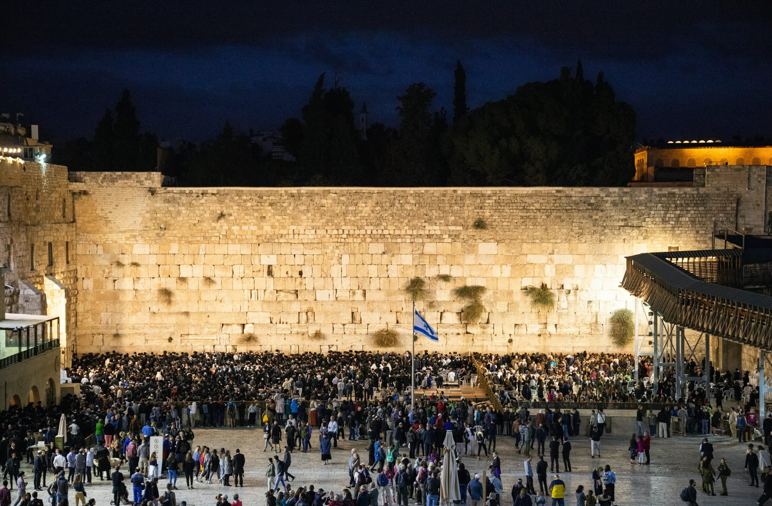 Vista a noite do Muro das Lamentações, em Jerusalém, com várias pessoas e uma bandeira do país no meio da multidão.