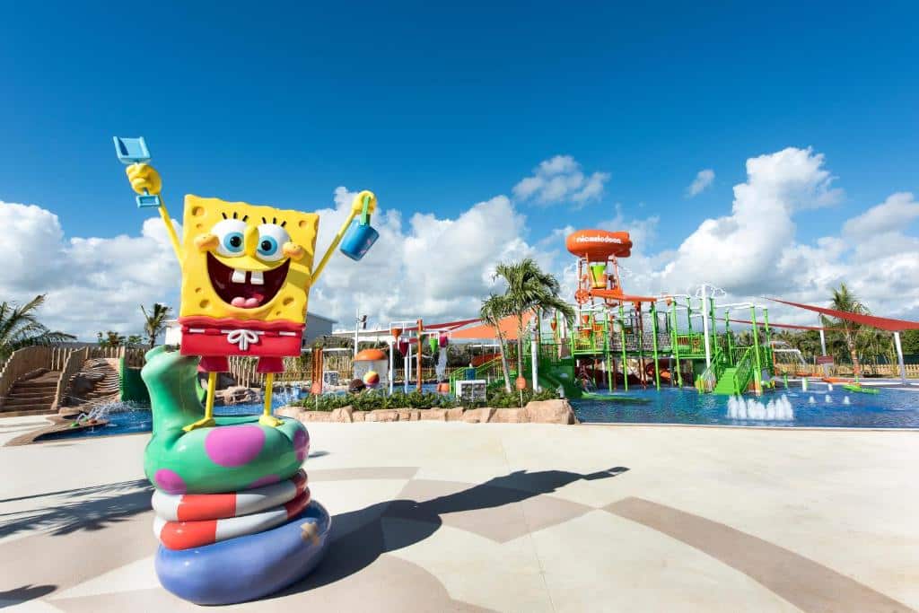 Escultura do Bob Esponja com as mãos para erguidas, em cima de várias bóias coloridas, com um parque aquáitico atrás. A imagem está de dia com bastante sol e serve para ilustrar o post como escolher um resort em Punta Cana