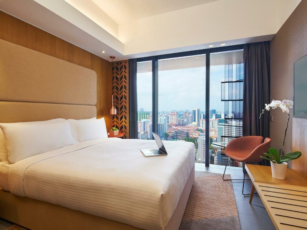 Quarto do Oasia Hotel Downtown, Singapore by Far East Hospitality. A cama de casal tem jogo de cama branco e um notebook aberto em cima. Há mesinhas laranja de cabeceira dos dois lados da cama com um vaso de planta em cima e um abajur na parede acima. Há um tapete bege embaixo da cama, e uma mesa com uma orquídea branca está de frente para a cama. Há uma televisão na parede acima, e uma potrona laranja está ao lado da mesa.
A parede da frente tem uma janela panorâmica com vista para a cidade, e há cortinas cinza abertas.