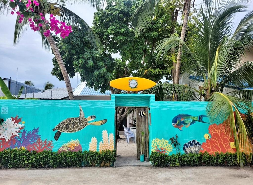 Frente do Ocean Breeze Maldives, um dos hotéis nas Maldivas, com um muro azul com desenho colorido de peixes no fundo do mar, uma prancha pequena em cima do portão de entrada e árvores no interior do local