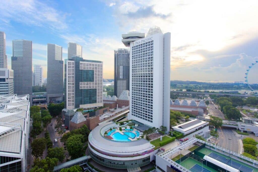 Visão de prédios ao redor do hotel Pan Pacific Singapore, uma das recomendações de onde ficar em Singapura. O hotel tem diversos andares e uma piscina na frente. A foto foi tirada de dia e o céu está azul.