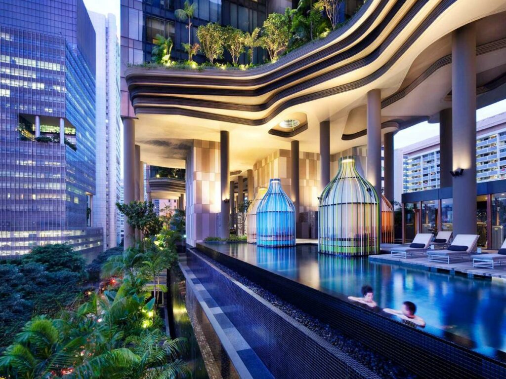 Piscina do hotel PARKROYAL COLLECTION Pickering, uma das recomendações de onde ficar em Singapura. A piscina é ao ar livre com visão da cidade à frente, e há árvores abaixo e acima da fachada moderna.. Espreguiçadeiras brancas estão atrás da piscina, onde duas pessoas estão paradas conversando.