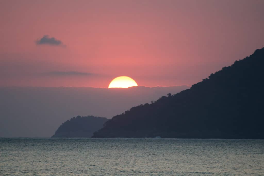 Pôr do sol na Praia do Paúba, São Sebastião, com mar a frente e ao fundo montanhas.