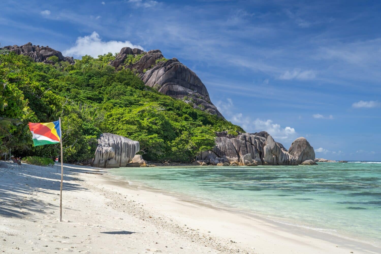 Uma praia com predas, mar verde claro, areia branca e uma bandeira de Seychelles
