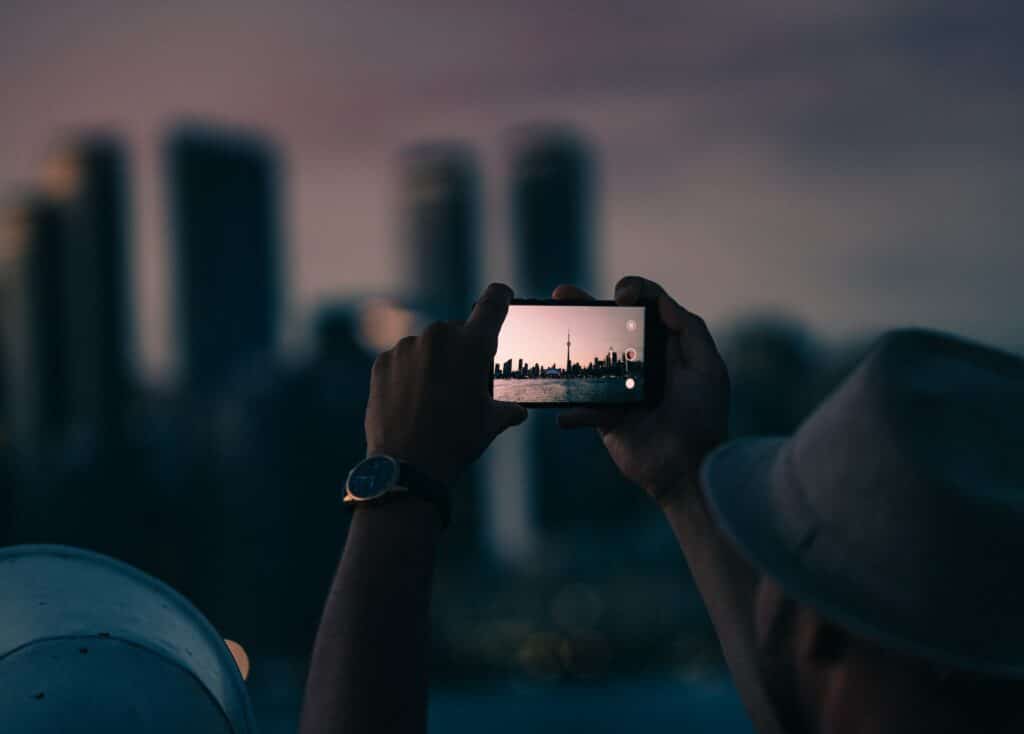 pessoa segurando um celular de costas, tirando uma foto da cidade de Toronto, no Canadá, no começo da noite, com vários prédios e um lago, para ilustrar o que você pode fazer utilizando o Passaporte Claro