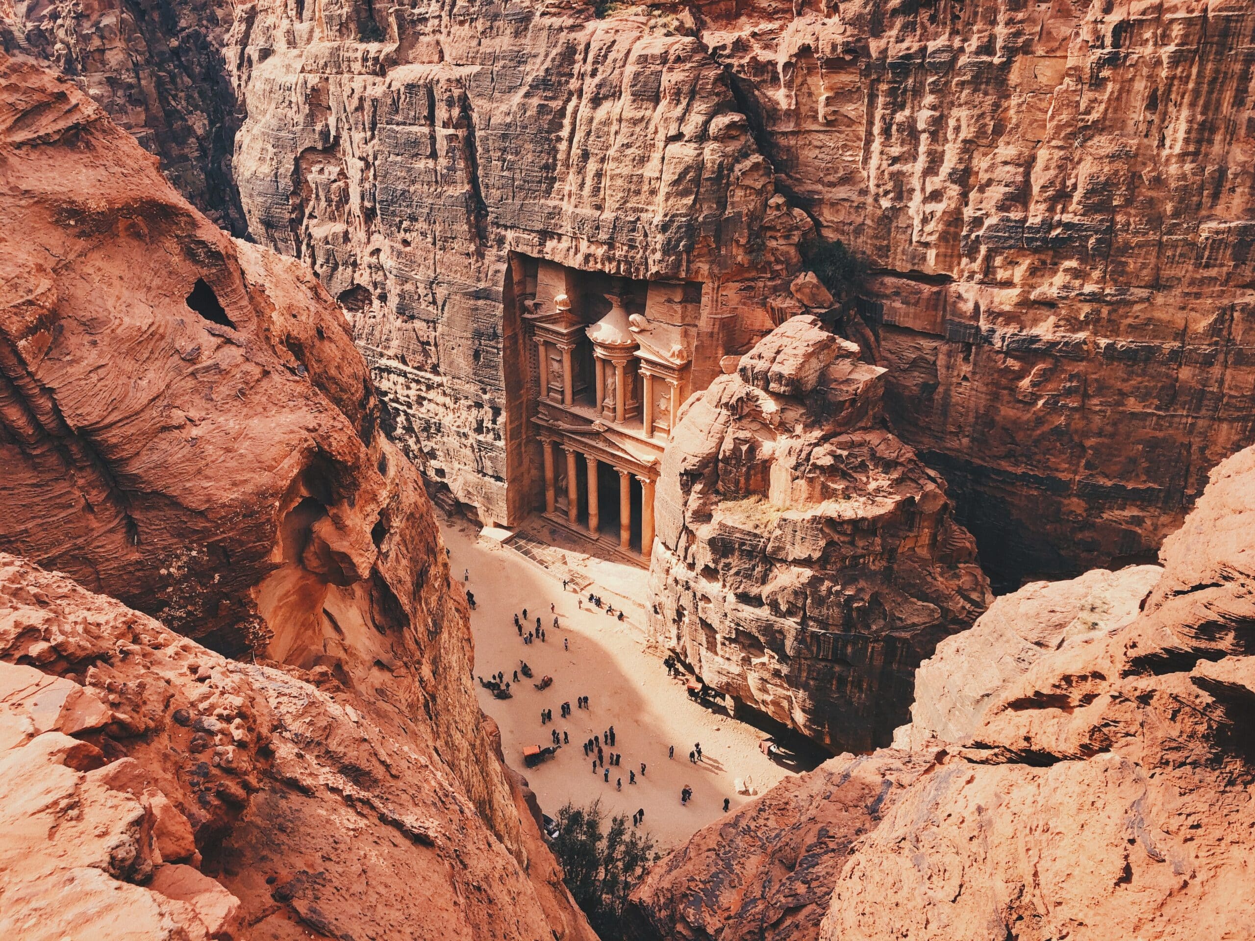 Vista de cima das ruína de Petra, na Jordânia, durante o dia em volta de montanhas rochosas em tons vermelho, com algumas pessoas abaixo. Representa chip celular Jordânia.
